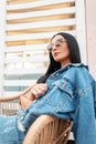 Pretty sexy young hipster woman in trendy sunglasses in a T-shirt in a vintage denim jacket in a blue skirt rest on a straw chair Royalty Free Stock Photo