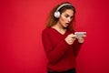 Pretty shocked young brunette curly woman wearing dark red sweater isolated on red backdrop wearing white headphones