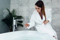 Pretty slim woman wearing bathrobe sitting on edge of bathtub filling up with water