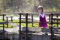 Pretty small blond long haired girl in nice red dress stands alone on old cement bridge leaning on wooden railings looking intent Royalty Free Stock Photo