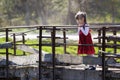 Pretty small blond long haired girl in nice red dress stands alone on old cement bridge leaning on wooden railings looking intent Royalty Free Stock Photo