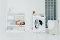 Pretty small child with pigtails lies on shelf of white console with neatly folded clean towels, poses in laundry room, pedigree Royalty Free Stock Photo