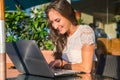 Pretty smiling blogger using laptop computer to write blogs while sitting at outdoor cafe on sunny day.