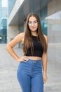 A pretty, smiling, curvy girl in glasses, black tank top, and blue jeans standing on the street