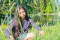 A pretty smiling girl with long hair crouched among meadow flowers in a clearing.