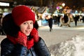 pretty smiling girl in warm clothes, wearing red hat, scarf and gloves is standing on street against background of winter New Year Royalty Free Stock Photo