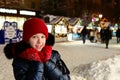 pretty smiling girl in warm clothes, wearing red hat, scarf and gloves stands against background of winter decorated street on New Royalty Free Stock Photo