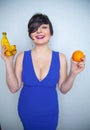 Cute chubby girl with short black hair in a blue dress holding an orange and bananas in her hands, smiling with love for fruits on