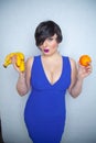 Cute chubby girl with short black hair in a blue dress holding an orange and bananas in her hands, smiling with love for fruits on