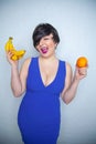 Cute chubby girl with short black hair in a blue dress holding an orange and bananas in her hands, smiling with love for fruits on