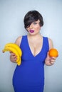 Cute chubby girl with short black hair in a blue dress holding an orange and bananas in her hands, smiling with love for fruits on