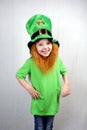 Pretty smiling littel girl in green clothes with decorative red beard in leprechaun\'s hat with gold clover leaf.