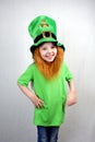 Pretty smiling littel girl in green clothes with decorative red beard in leprechaun\'s hat with gold clover leaf