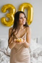 Pretty smiling woman with long hair lies in bed with a cake on a plate and a candle and celebrates her birthday Royalty Free Stock Photo