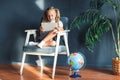 Pretty smiling blondy girl relaxing on a chair near the globe indoors at home with a tablet pc in her socks and jeans