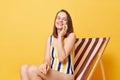 Pretty smiling woman wearing striped swimming suit sitting on deck chair isolated over yellow background talking on smart phone Royalty Free Stock Photo