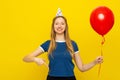 Pretty smiling young woman in a birthday cap is pointing finger down, holds red inflated helium balloon, celebrates birthday or Royalty Free Stock Photo