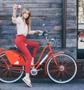 Pretty smiling young woman using taking self portrait on pink vintage camera with retro bicycle over old wood planks background