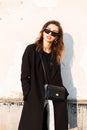 Pretty stylish hipster young woman in dark sunglasses in a vintage long coat in white jeans with a black handbag stands