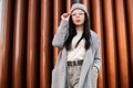 Pretty stylish young hipster woman in fashionable coat in knitted gray hat in vintage glasses posing near metal wall in the city. Royalty Free Stock Photo
