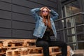 Pretty stylish young hipster woman in purple trendy glasses in blue stylish denim jacket in black vintage jeans posing on wooden Royalty Free Stock Photo