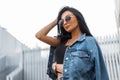 Pretty stylish young hipster woman in a trendy black t-shirt in sunglasses in a vintage denim jacket posing outdoors Royalty Free Stock Photo