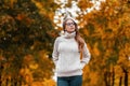 Pretty stylish young hipster woman in a trendy white warm knitted sweater in blue jeans in fashionable glasses posing in the park Royalty Free Stock Photo