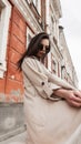 Pretty stylish young woman in fashionable sunglasses in beige spring trendy raincoat whirls on street near vintage building. Royalty Free Stock Photo