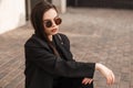 Pretty stylish young woman in fashionable youth black clothes in sunglasses sit on vintage tile in city. American girl fashion Royalty Free Stock Photo