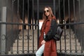 Pretty stylish young woman model in brown fashion trench coat in sunglasses with leather backpack posing near iron gate on street Royalty Free Stock Photo