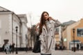 Pretty stylish young woman model in a light fashionable long trench coat with a leather trendy bag stands on a city street on a