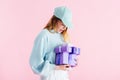 Pretty teenage girl in cap looking at purple gifts isolated on pink