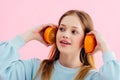 Pretty teenage girl holding orange halves near ears isolated on pink