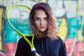 Pretty Teenage Tennis Player looking at you camera holding tennis racket outdoors graffiti background