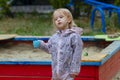 Pretty toddler girl wearing rain jacket playing with blue cup near the sandbox on the playground