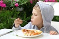 Pretty toddler is holding fork with a slice of pizza.The look of pleasure on his face.Beautiful floowers and summer foliage n the