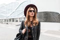 Pretty urban hipster young woman in a fashionable hat in stylish sunglasses in a vintage leather jacket with a backpack walks Royalty Free Stock Photo