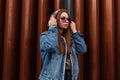 Pretty urban young hipster woman puts on a hood. Beautiful girl model in stylish denim jacket with glamorous purple glasses posing Royalty Free Stock Photo