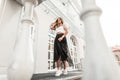 Pretty urban young woman in an elegant black long skirt in a trendy blouse in leather sneakers posing near a vintage building in