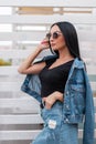 Pretty urban young woman fashion model with luxurious black hair in trendy blue denim clothes in stylish dark sunglasses Royalty Free Stock Photo