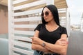 Pretty urban young woman fashion model with luxurious black hair in trendy black t-shirt in stylish dark sunglasses poses outdoors