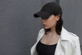 Pretty urban young woman in a fashionable black baseball cap in a trendy white leather jacket in a t-shirt near the gray wall Royalty Free Stock Photo