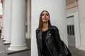 Pretty urban young woman in a fashionable black leather jacket in an elegant dress with a stylish bag stands near a vintage white