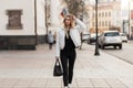 Pretty urban young woman in a fashionable white jacket in vintage black jeans with a black stylish leather bag on the street.