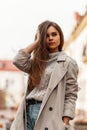 Pretty urban young woman straightens hair. Beautiful stylish girl a fashion model in a fashionable trench coat in a vintage gray