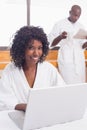 Pretty woman in bathrobe using laptop at table with partner in background Royalty Free Stock Photo