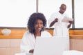 Pretty woman in bathrobe using laptop at table with partner in background Royalty Free Stock Photo