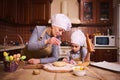 Pretty woman in chef& x27;s hat and beige apron, flouring dough, preparing Easter cake with her daughter in the home