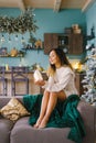 Pretty woman is sitting on the sofa near the Christmas tree and holding a toy in her hands a glowing cozy house in anticipation of