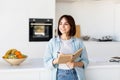 Pretty woman taking notes, holding notepad and pen, standing in kitchen interior and looking aside, copy space Royalty Free Stock Photo